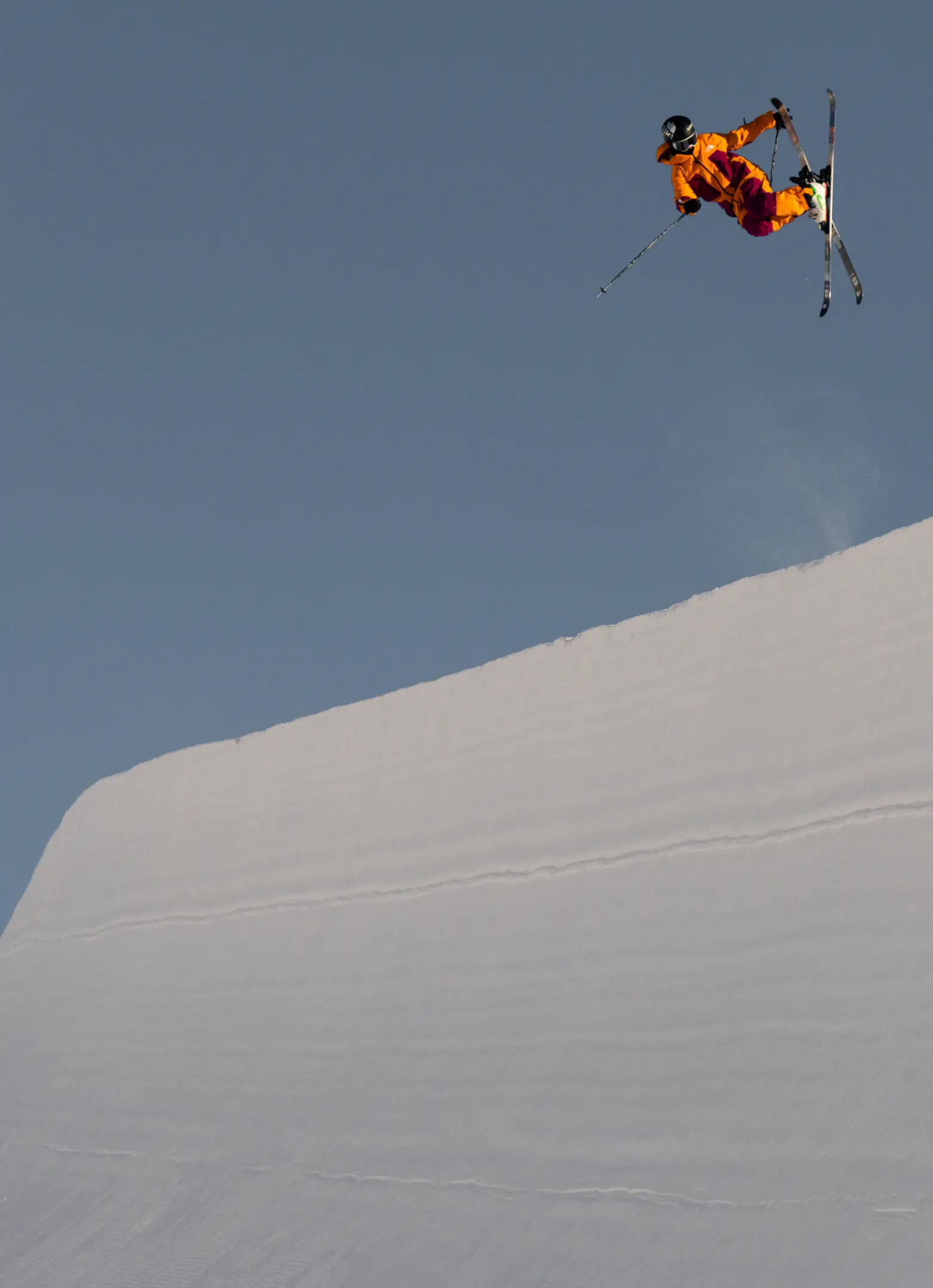 A freestyle skier in a bright orange jacket crosses their skis mid-air above the halfpipe lip against a clear blue sky.