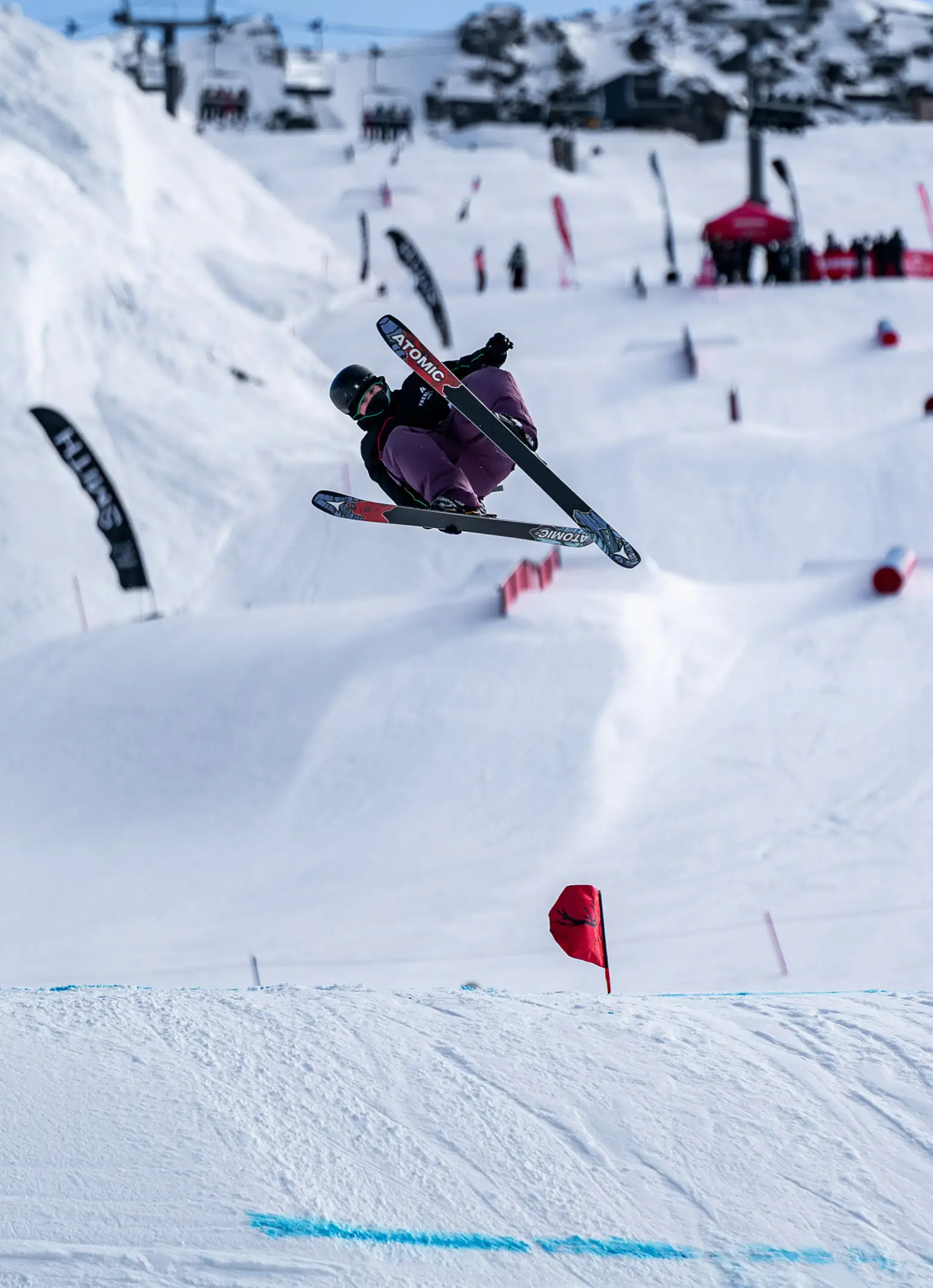 A freestyle skier performs an aerial trick in a terrain park, grabbing their Atomic skis mid-air above a series of snow features, with red flags and spectators visible in the background.