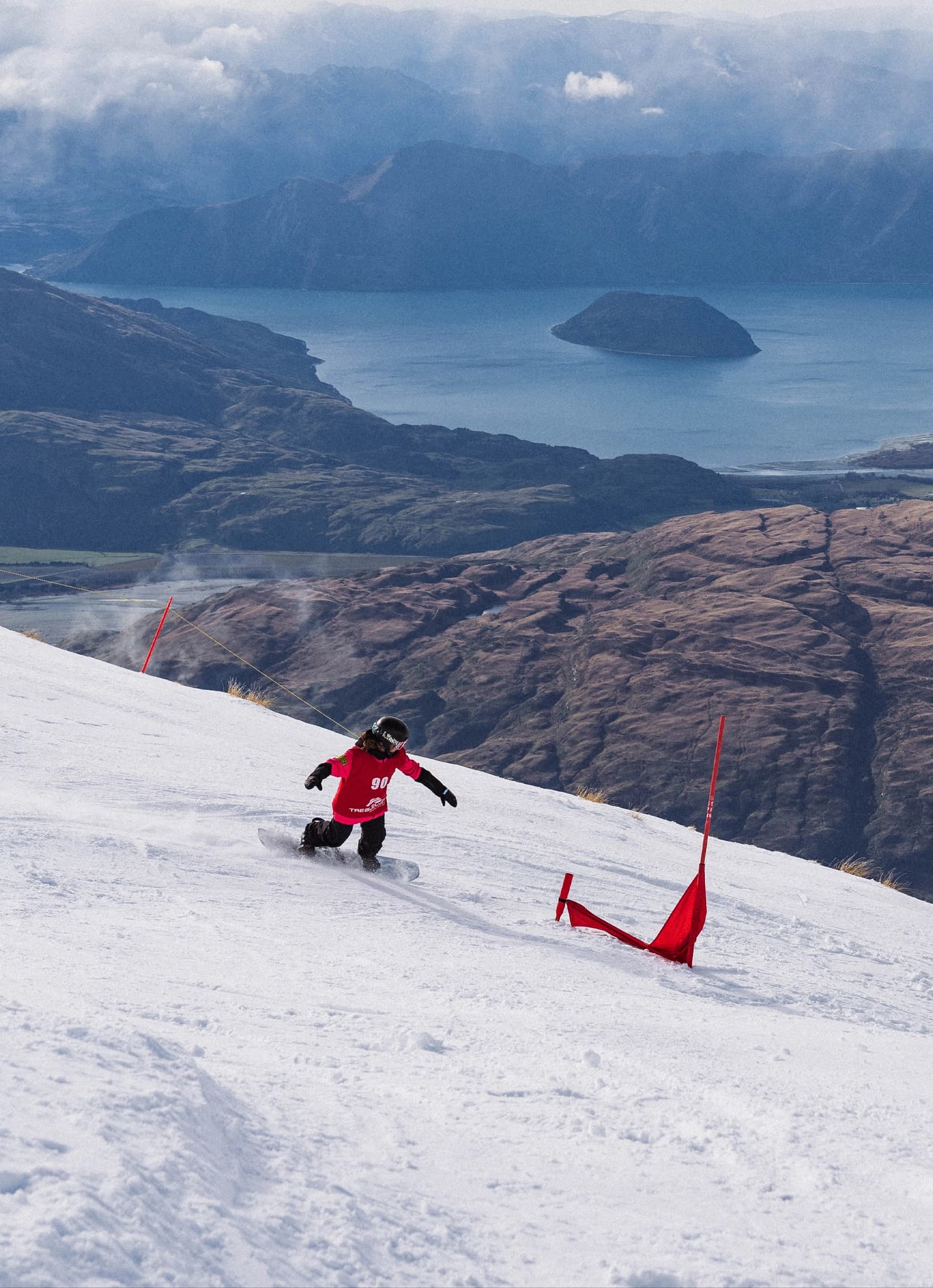 Snowboarder racing through a banked slalom course with sweeping lake views below.