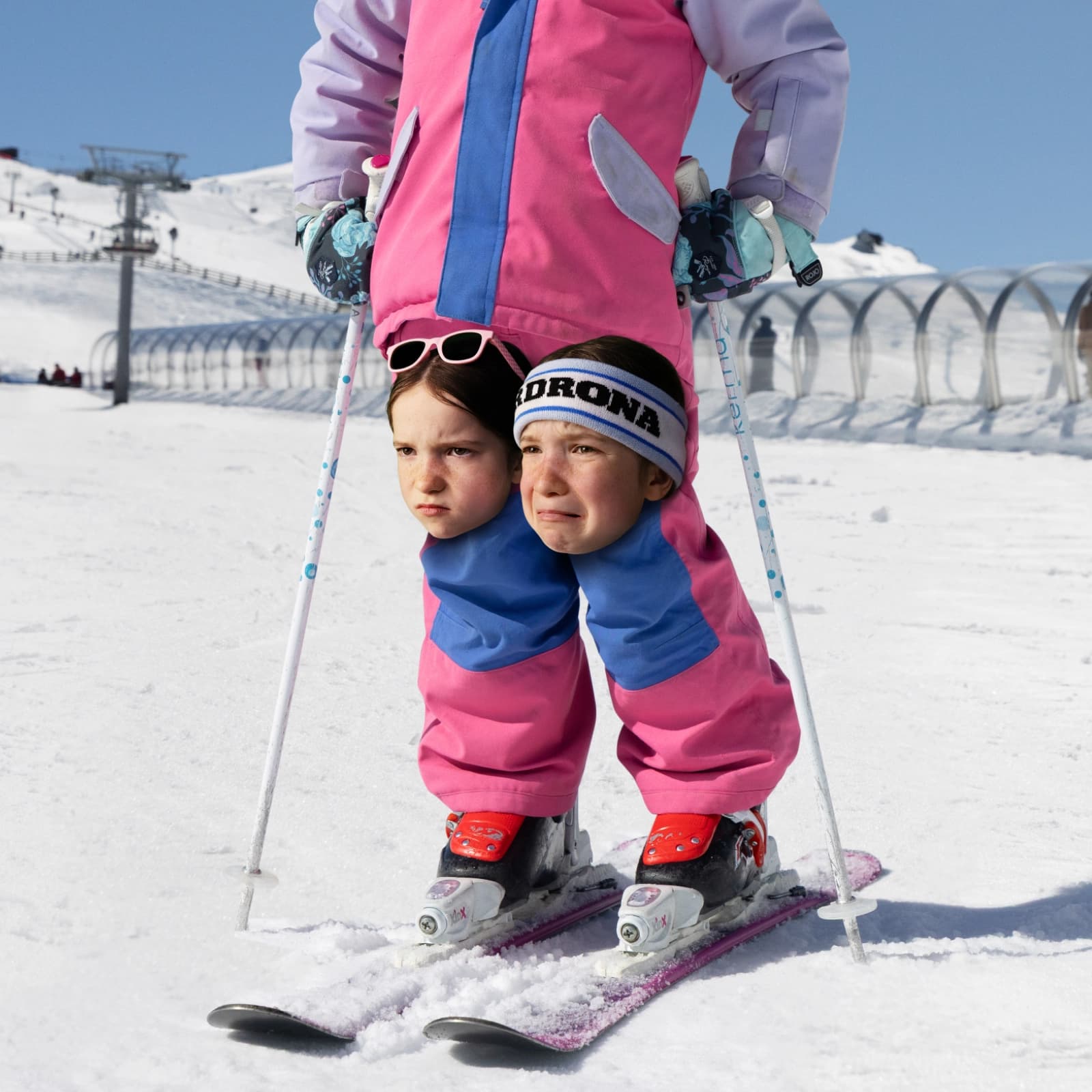 Surreal scene of a skier on a snowy slope wearing a pink suit, with two children’s heads appearing on the skiers knees.