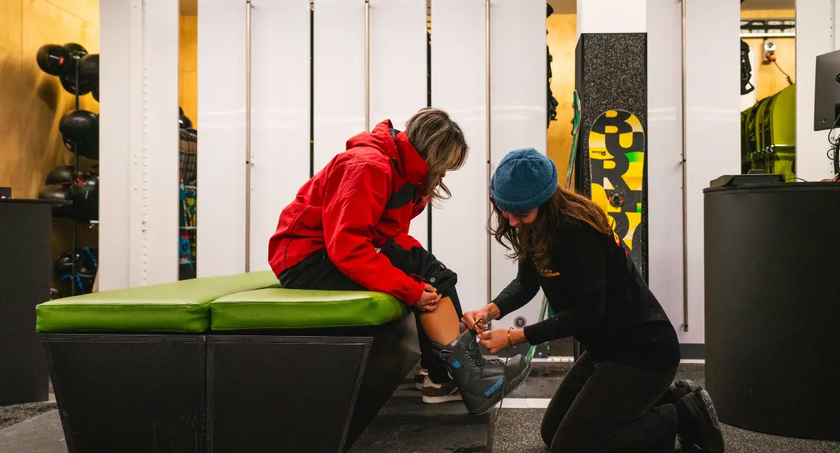 A staff member kneels to help a guest try on snowboard boots in a modern ski and snowboard rental shop.