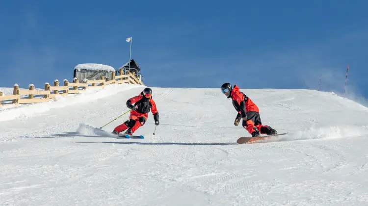 A skier and a snowboarder in red jackets carving down a groomed snowy slope under a clear blue sky, with a building at the top of the hill.