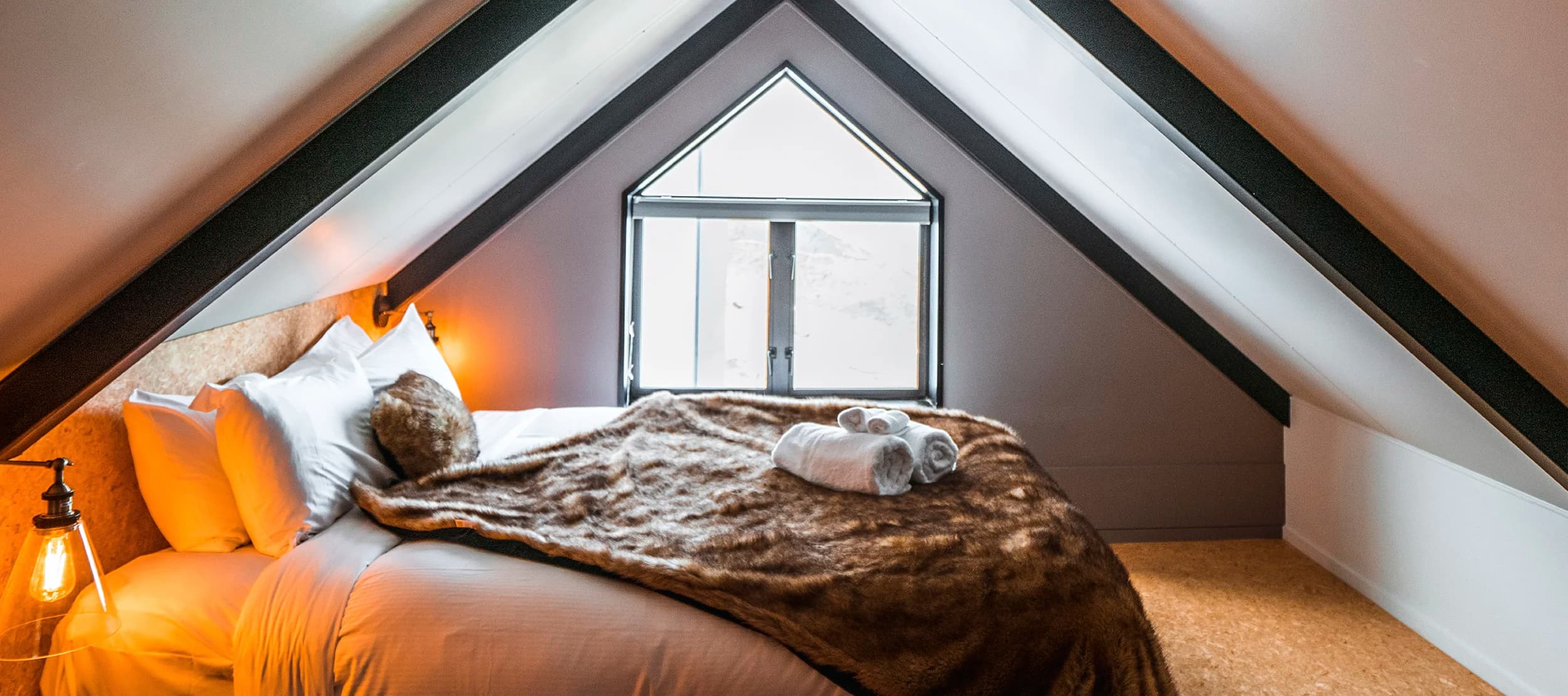 A cozy attic bedroom featuring a sloped ceiling with exposed beams, a comfortable bed draped with a soft fur blanket, and warm lighting, creating an inviting atmosphere at the Cardrona Apartments.
