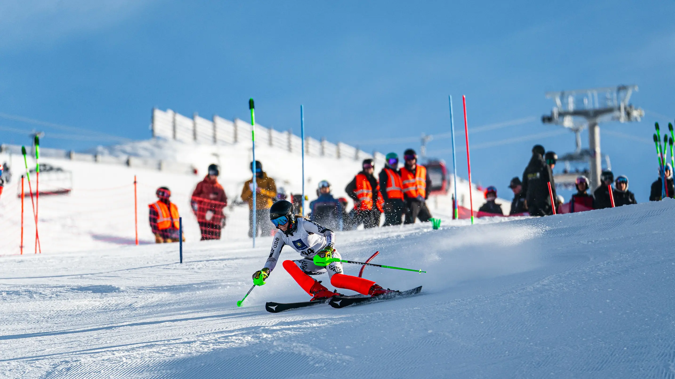 A female slalom ski racer in a white and red suit carves aggressively through a gate, with orange-vested officials and spectators watching from the sideline.