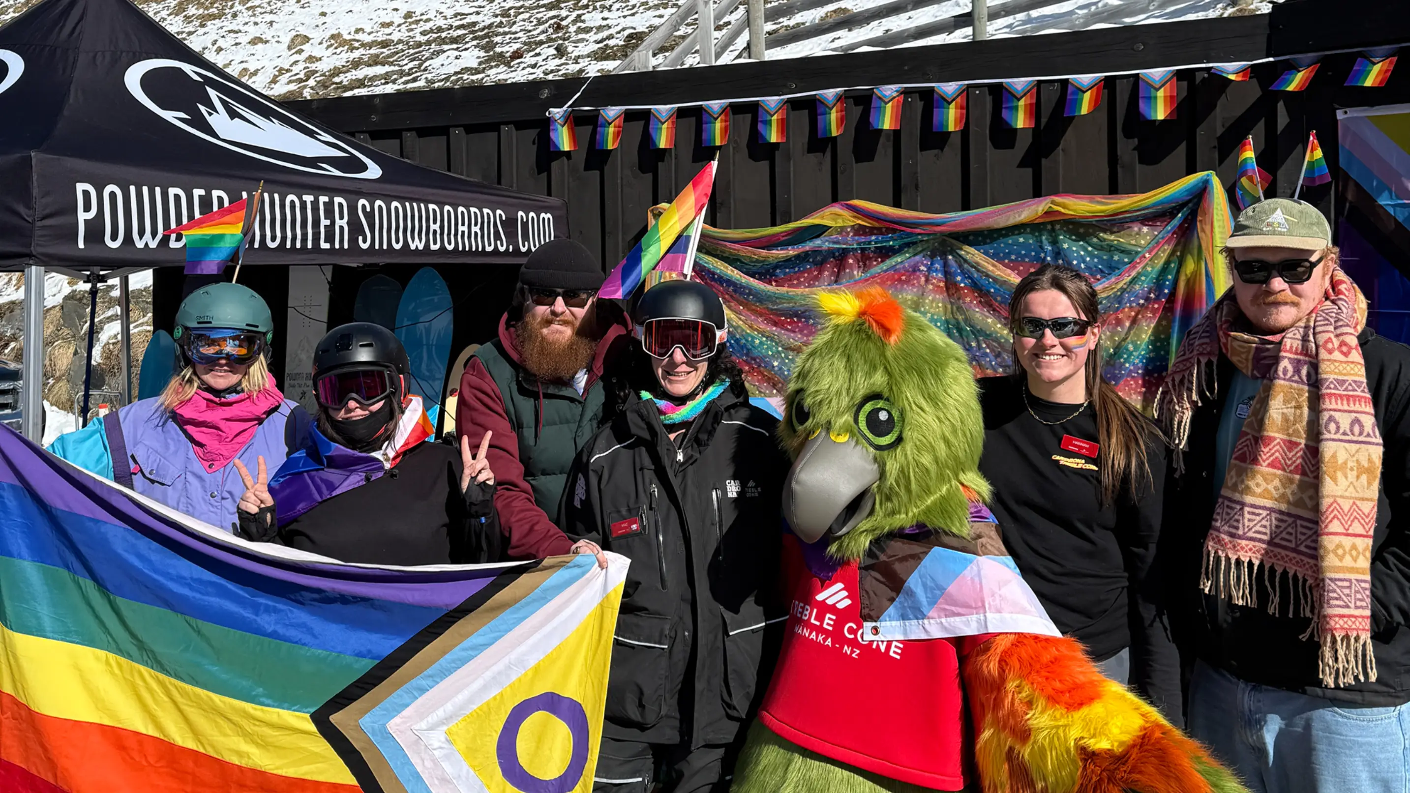A kea bird mascot stands with a group of smiling people on a ski field, while holding a pride flag.