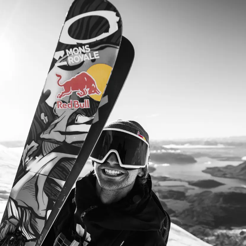 Black and white headshot of a skier on the smiling on the mountain holding his skis, which have Mons Royale and Red Bull stickers on them.