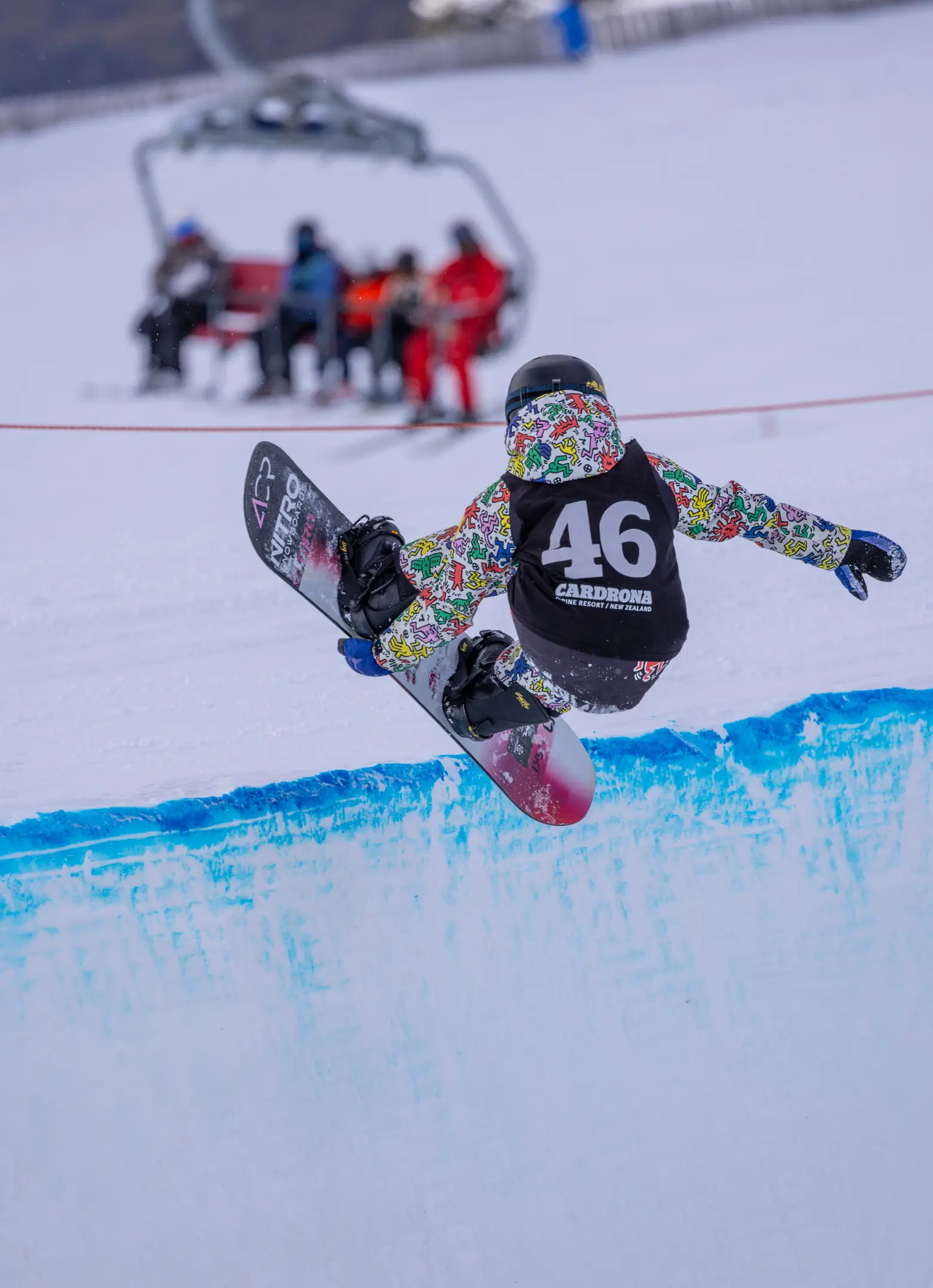 A snowboarder wearing bib 46 catches air out of a halfpipe at Cardrona Alpine Resort, with a chairlift and riders visible in the background.