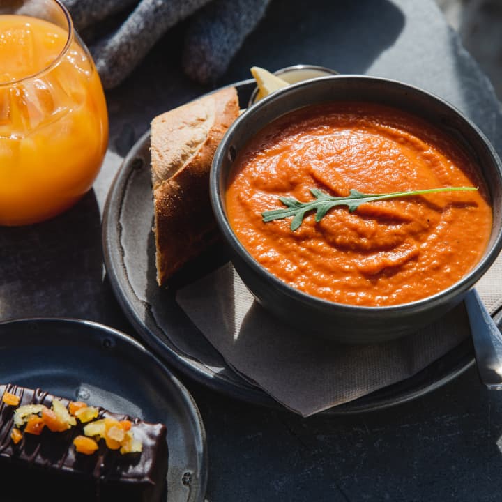 A bowl of roasted tomato soup with toast and a chocolate slice on dark plates, with a glass of orange juice.