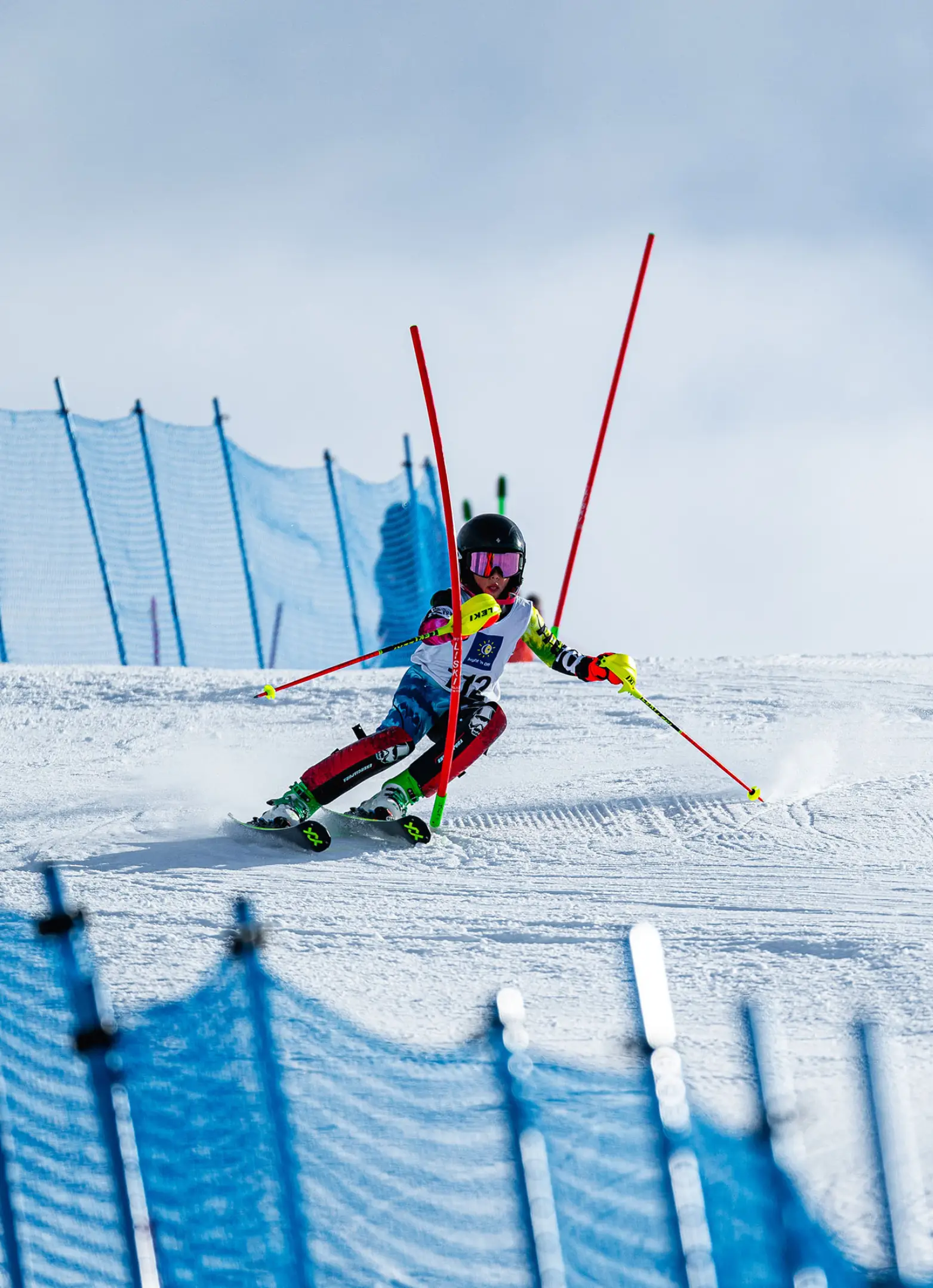 A young junior alpine ski racer in a colourful race suit navigating a slalom course, carving past red and blue gates on a groomed snow slope.