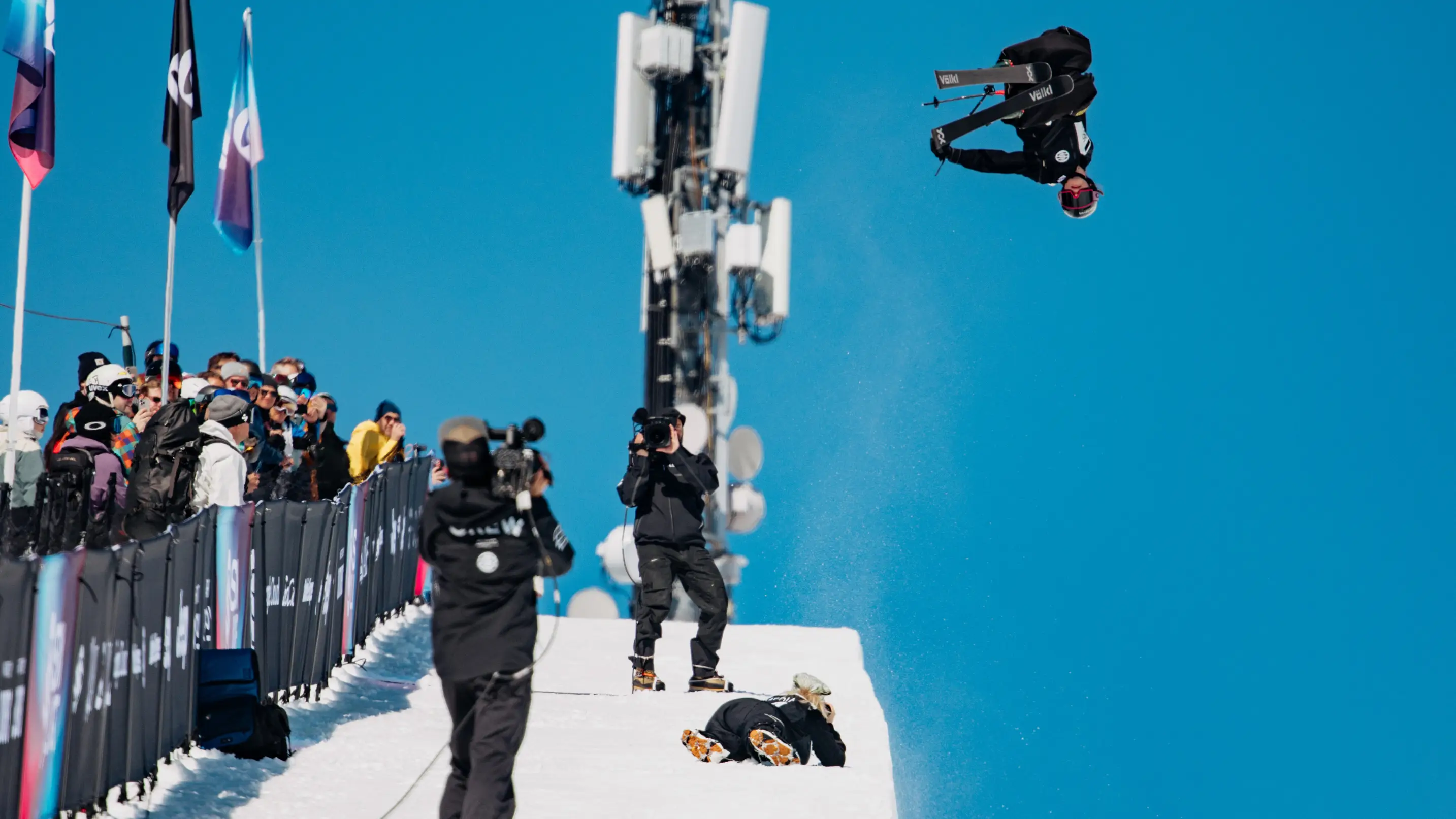 A freestyle skier performs an inverted aerial trick above a snowy competition course as spectators and photographers watch.