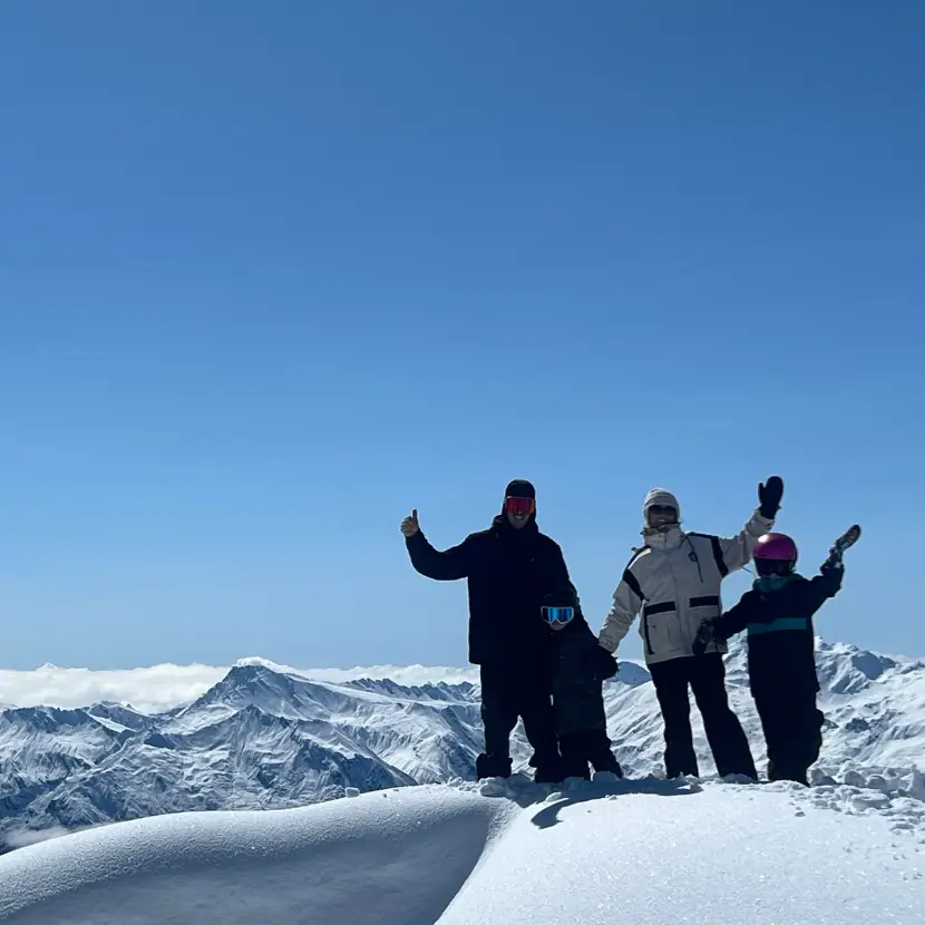 A family of skier or snowboarders stand at the top of a snowy mountain on a bluebird day with their arms in the air.