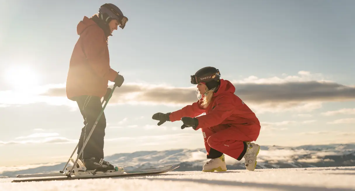 Ski instructor helping a beginner adjust skis on a snowy slope at sunset, with mountains in the background.