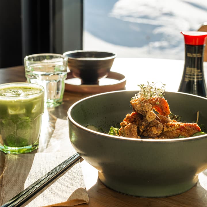 A bowl of crispy fried chicken over greens beside an iced matcha drink, with chopsticks and soy sauce on a sunlit table.