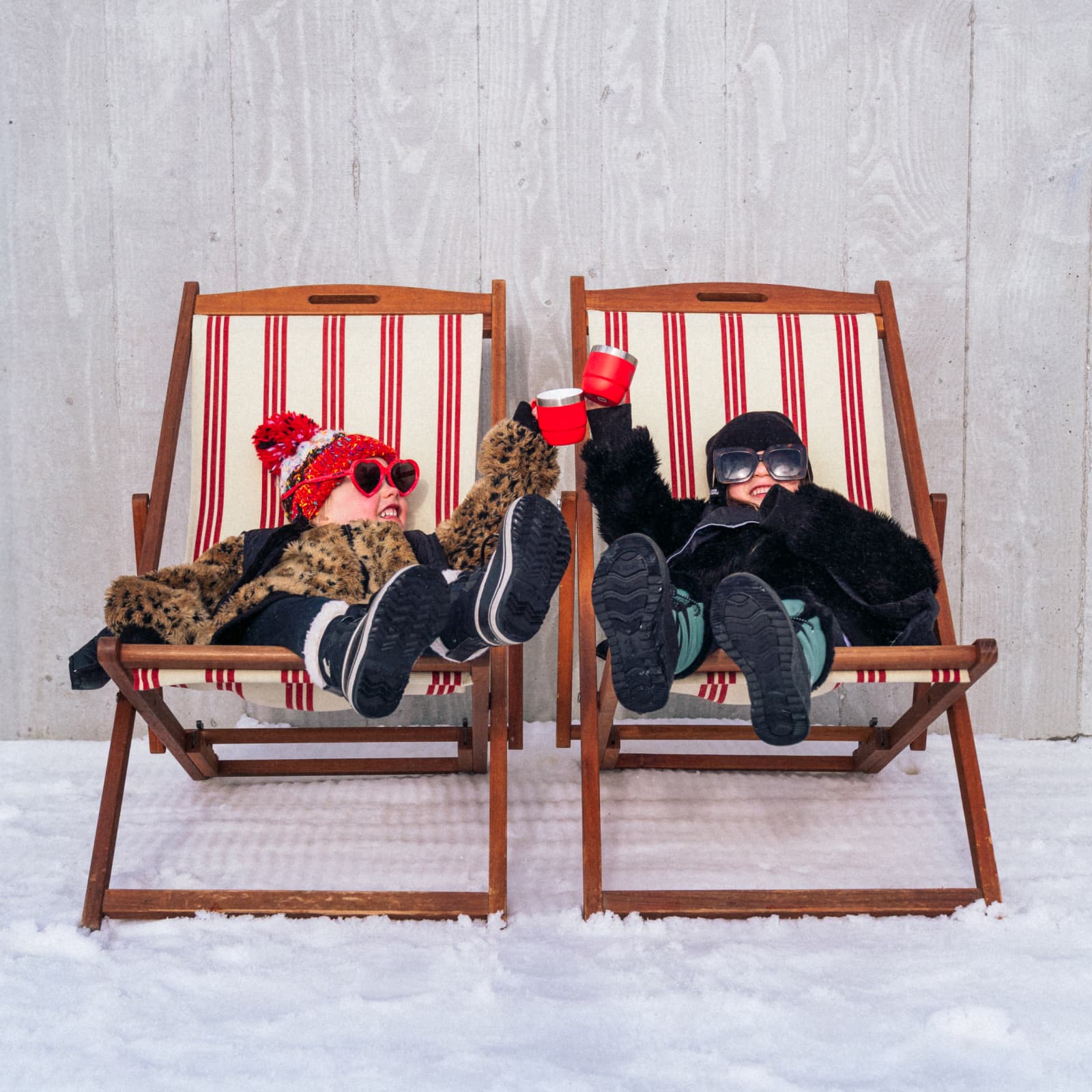 Two kids cheers while sitting on deck chairs on a snowy mountain, dressed in ski gear.