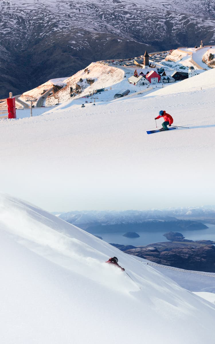 Skiers descend snowy slopes above an alpine lake and mountain base area.