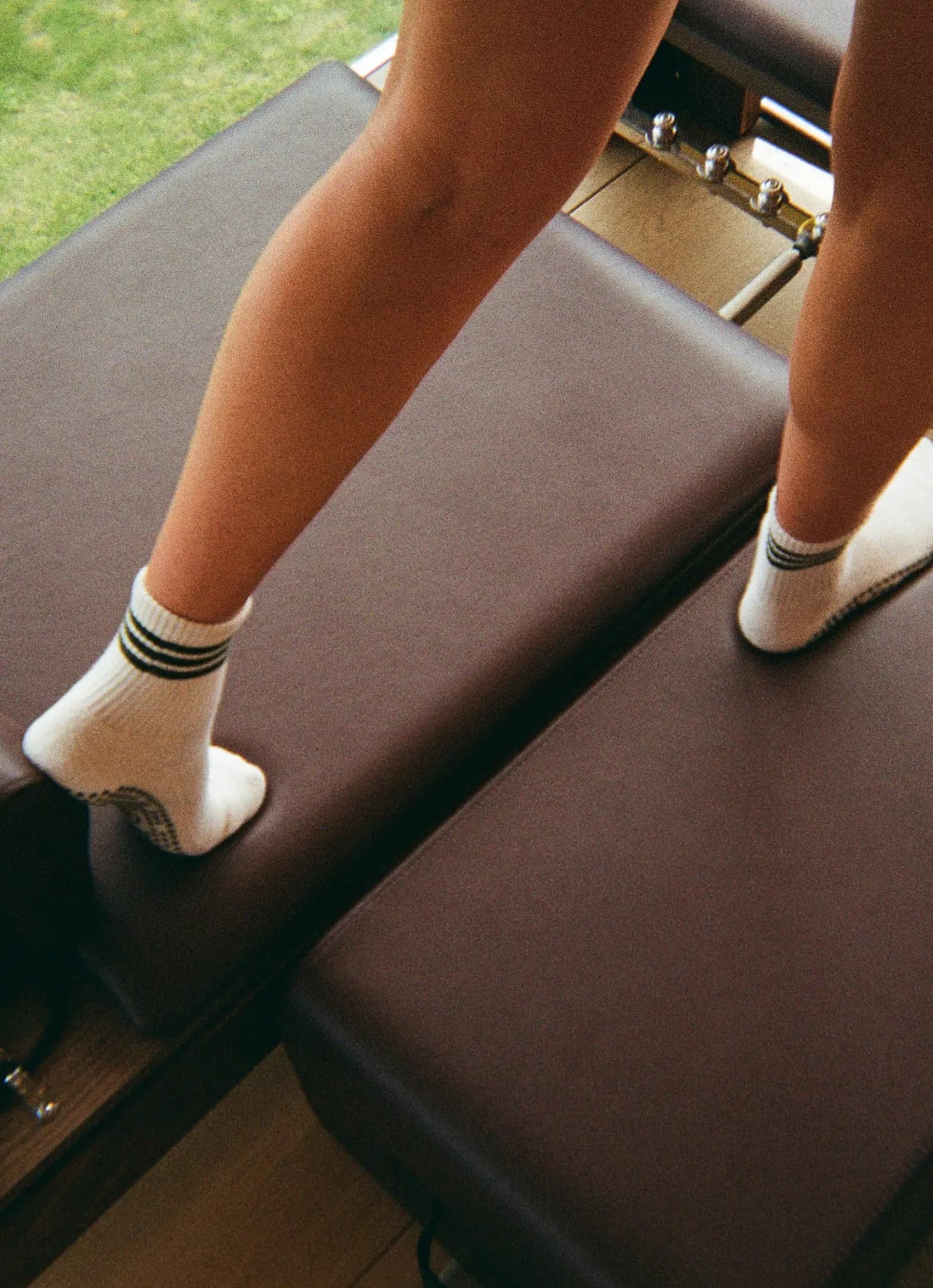 A person standing on a Pilates reformer machine in white grip socks, with green grass visible through the window behind them.