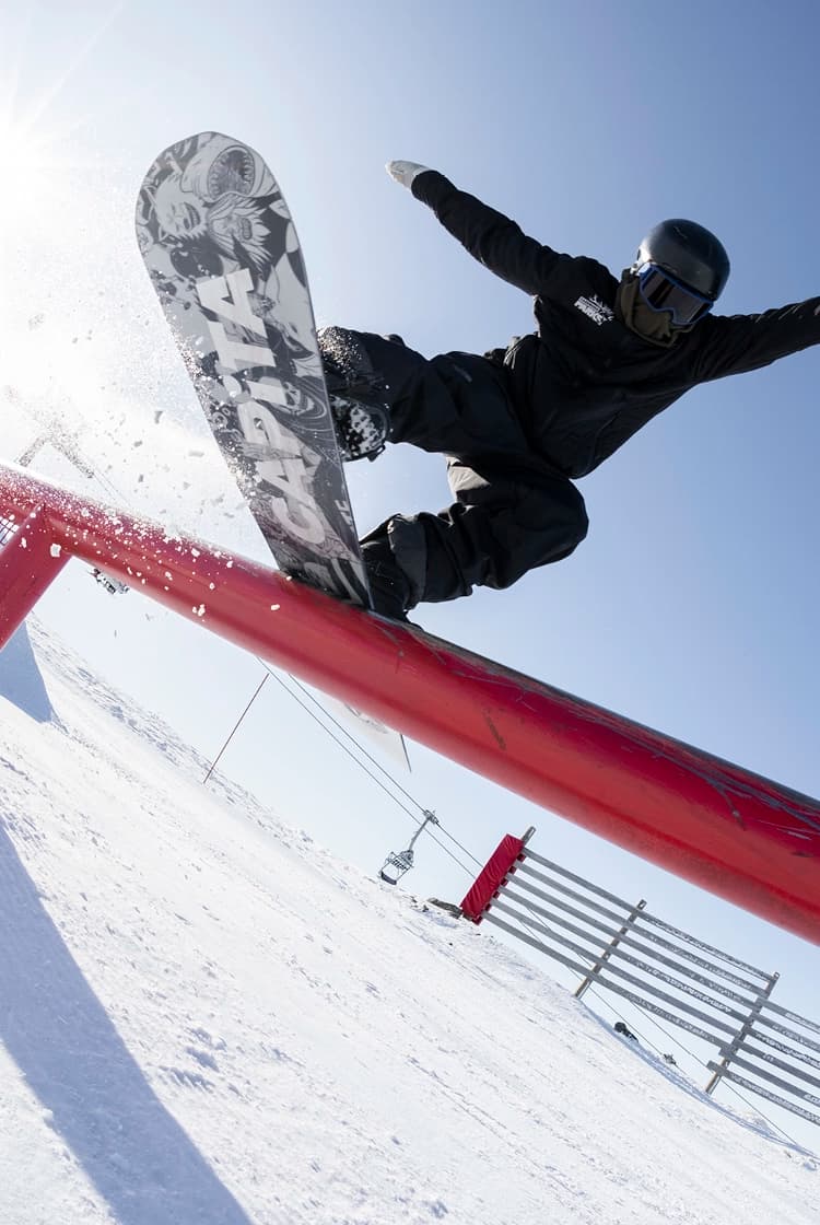 A snowboarder performing a trick while airborne over a bright red rail in a snow-covered terrain park. The rider is in mid-motion, board vertical to the ground, showcasing the artwork on the underside of the board. There is a snowy mountain landscape in the background, on a clear blue day, along with other park features and a few spectators.
