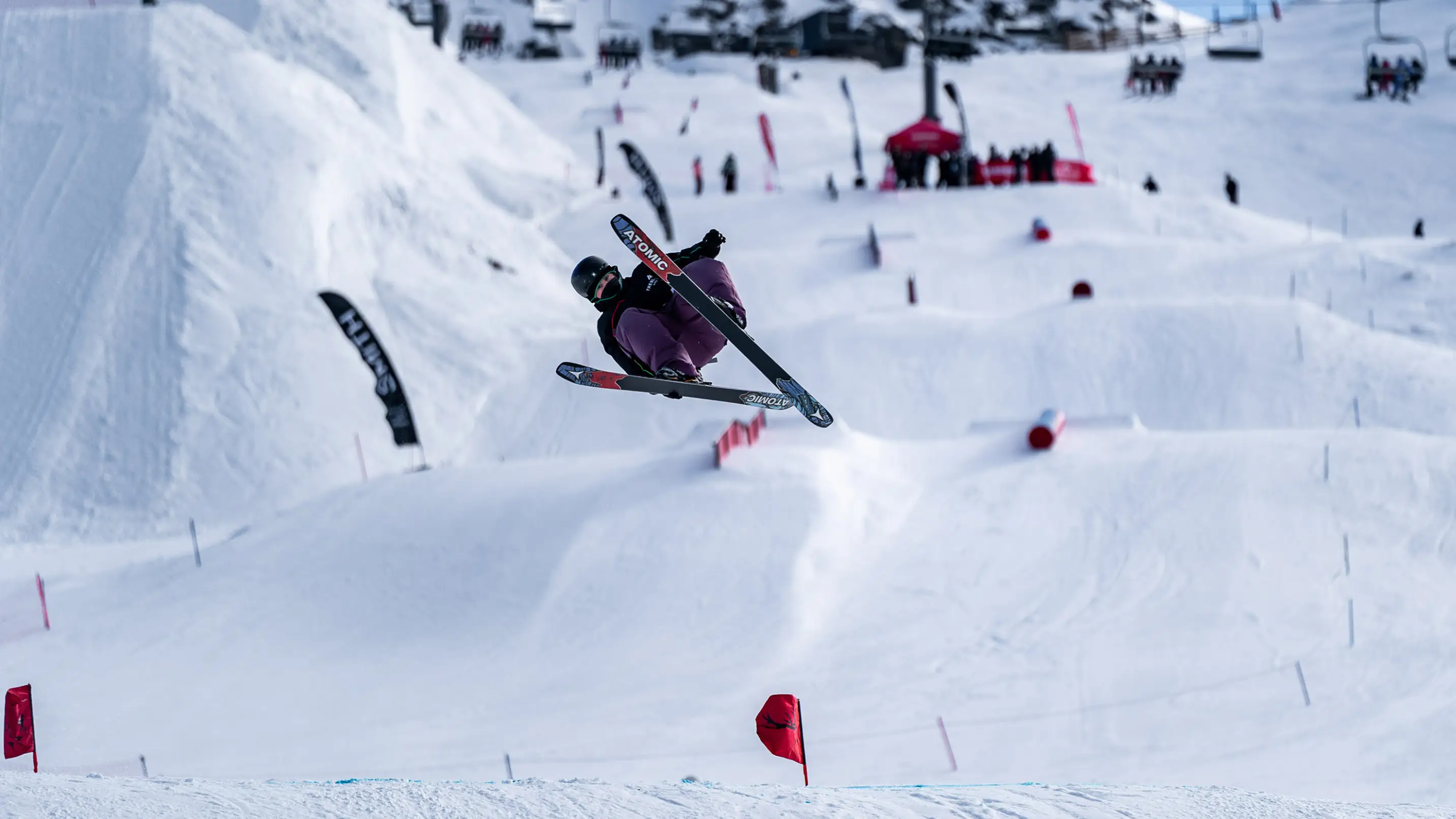 A freestyle skier performs an aerial trick in a terrain park, grabbing their Atomic skis mid-air above a series of snow features, with red flags and spectators visible in the background.