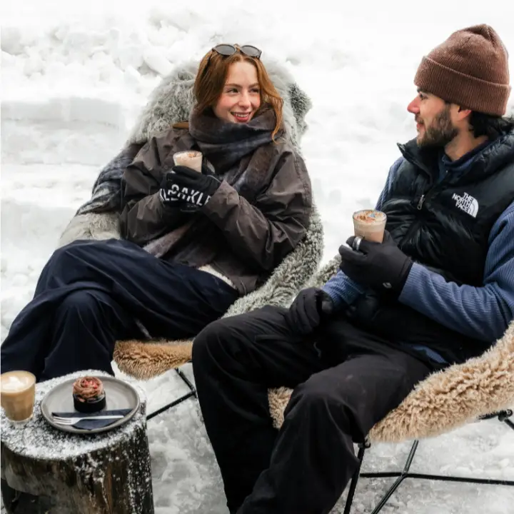 Two people enjoying hot drinks while relaxing in chairs surrounded by snow.