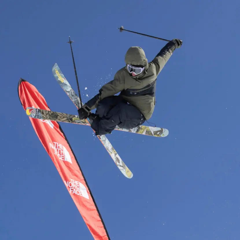 A skier in the air with his skis crossed doing a grab. There are blue skies and a red flag in the background.
