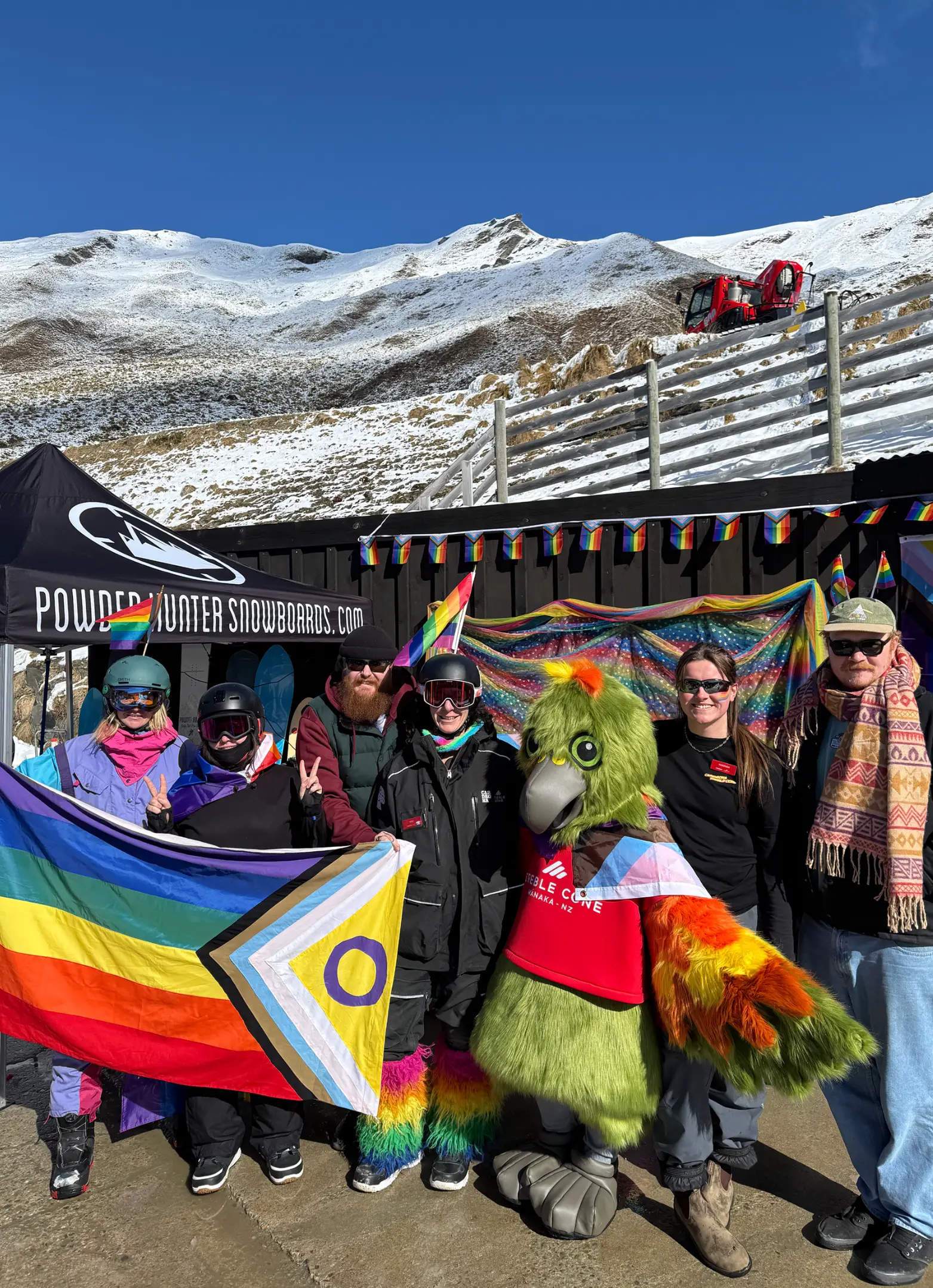 A kea bird mascot stands with a group of smiling people on a ski field, while holding a pride flag.