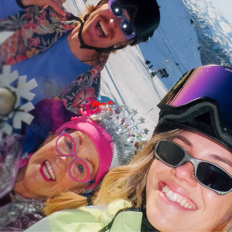 Selfie of two smiling skiers with a fairy god mother, all dressed in bright colours, on a snowy mountain on a sunny day.