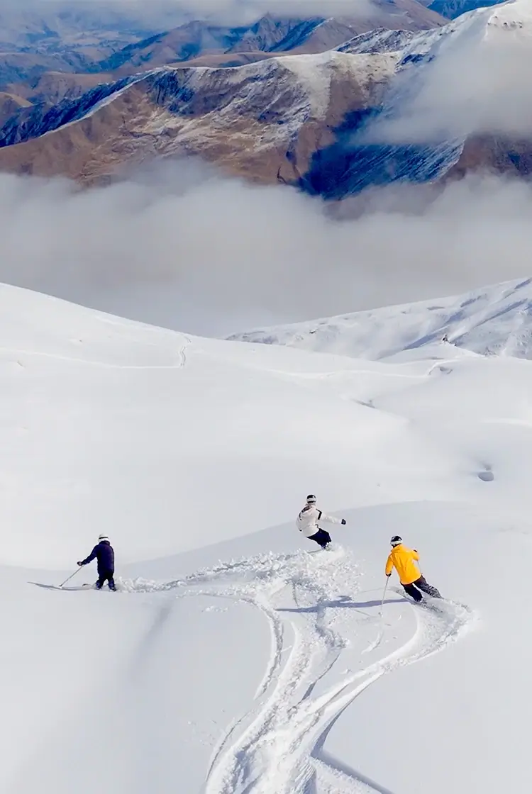 Two skiers and a snowboarder descend a wide, untouched powder slope at Cardrona, surrounded by snow-covered peaks and dramatic alpine scenery under a cloud-filled sky.