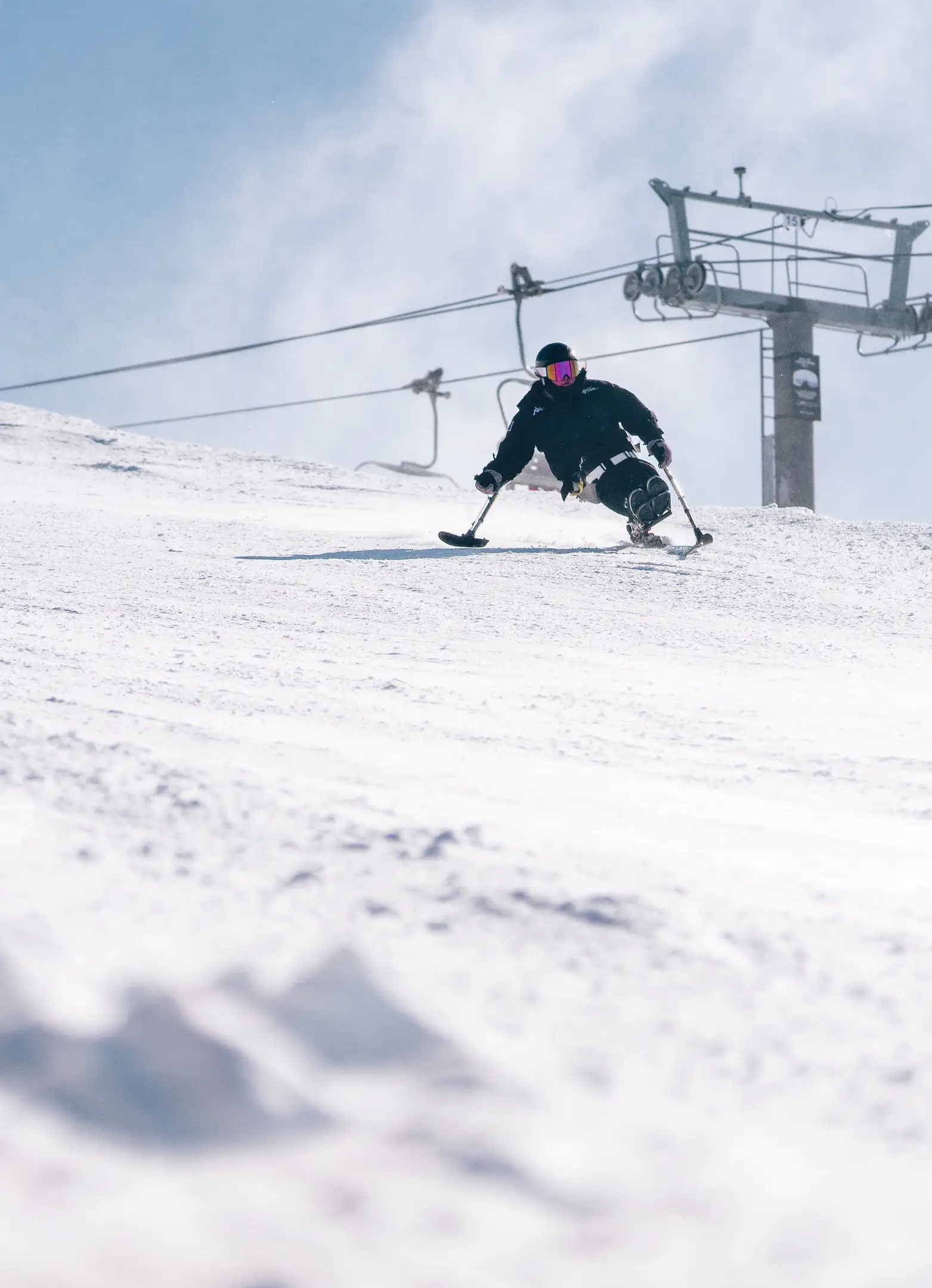Adaptive sit-skier carving down a snowy slope with a chairlift in the background on a bright winter day.