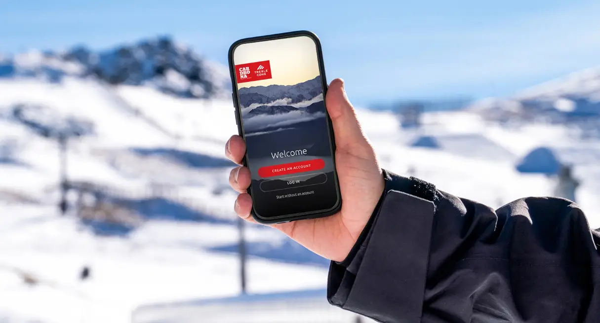 A hand holding a smartphone displaying the Cardrona and Treble Cone ski resort app welcome screen, with a snowy mountain landscape in the background.