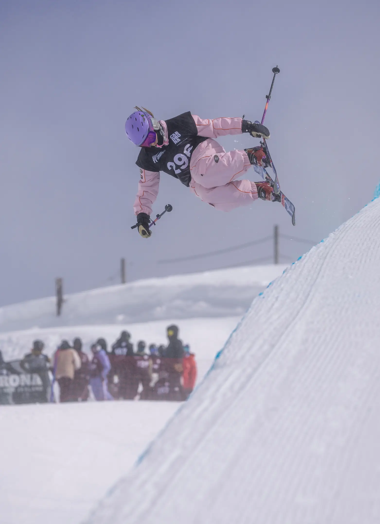A freeskier wearing bib 29, in a pink outfit and purple helmet, grabs their ski mid-air above a halfpipe at Cardrona, with spectators watching below.