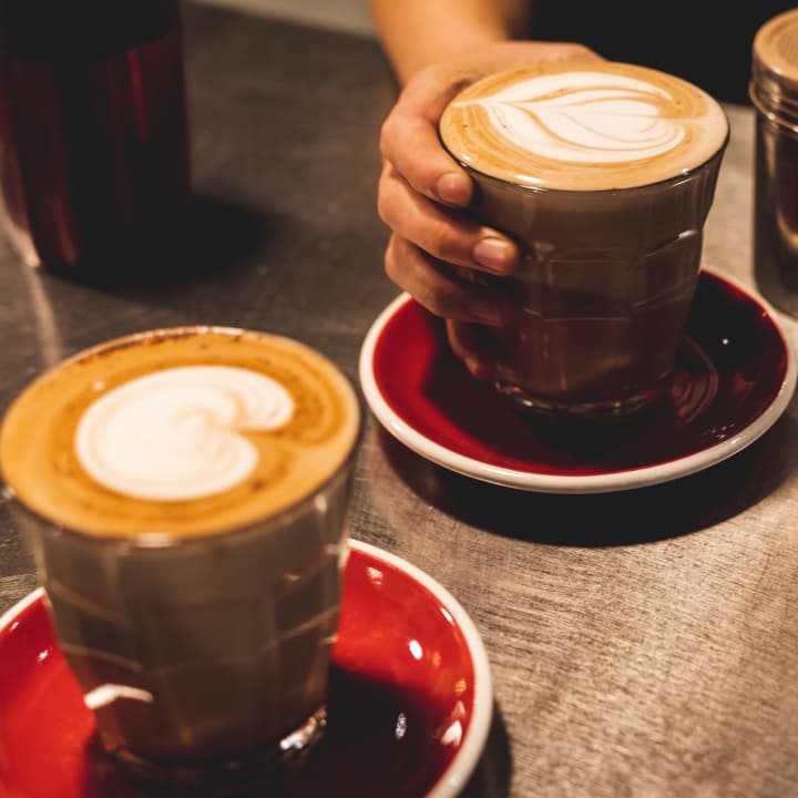 Two latte art coffees in glass cups on red saucers, one being handed across a counter.
