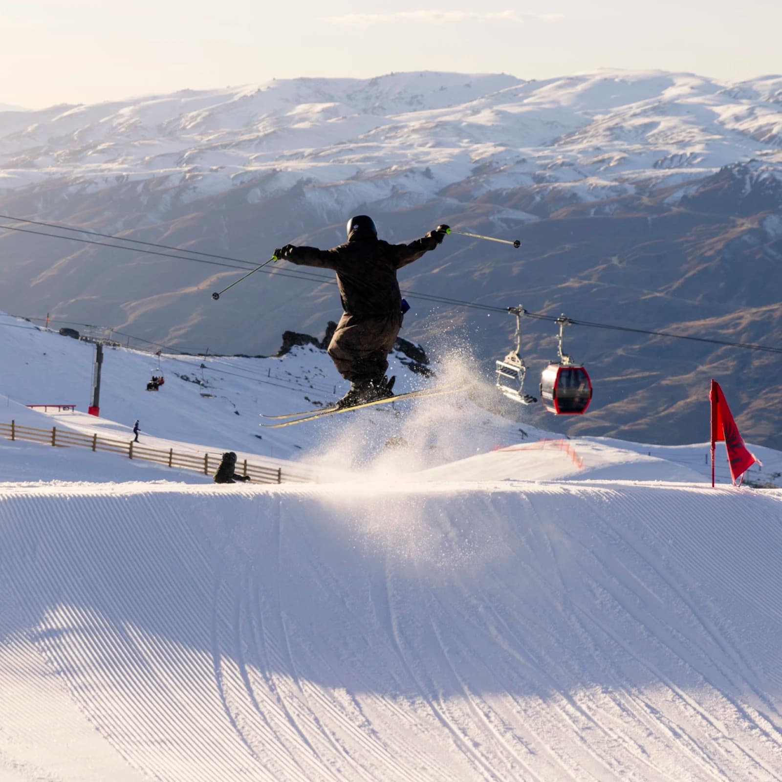 A skier performing a jump on at a snow covered terrain park on a clear day. A snowy mountain landscape can be seen in the background, along with a chair lift with a red gondola cabin.