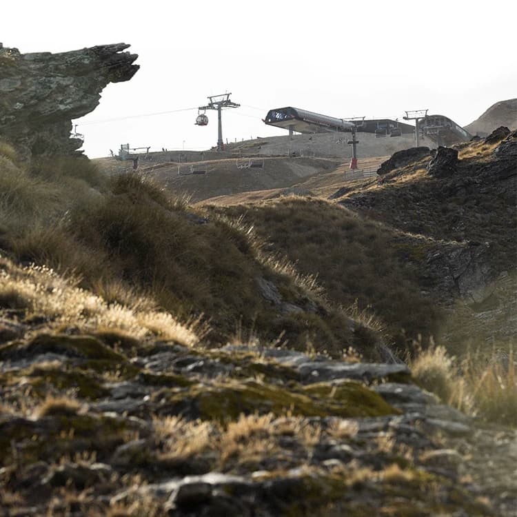 A rugged mountainous landscape with steep rock formations and grassy slopes during summer. A chair lift, with gondolas and chairlifts, is visible in the background.