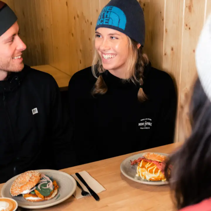 A woman in a beanie laughs with a friend over coffee and breakfast sandwiches at a café table.