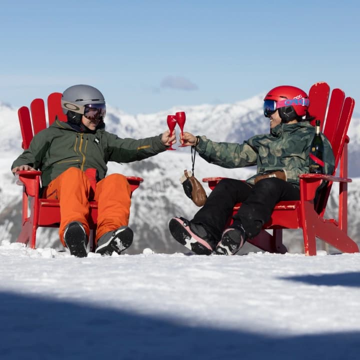 Two skiers in red Adirondack chairs on the snow toast with red wine glasses against a mountain backdrop.