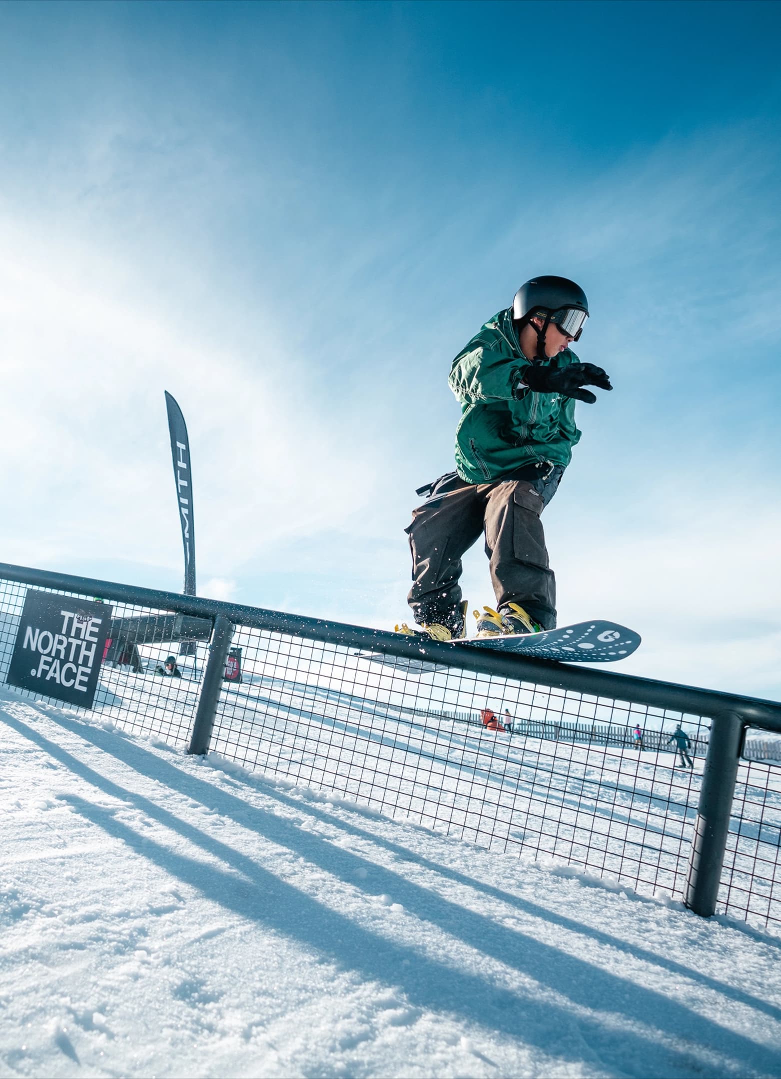 A snowboarder rides a North Face rail feature with flags and blue skies behind.