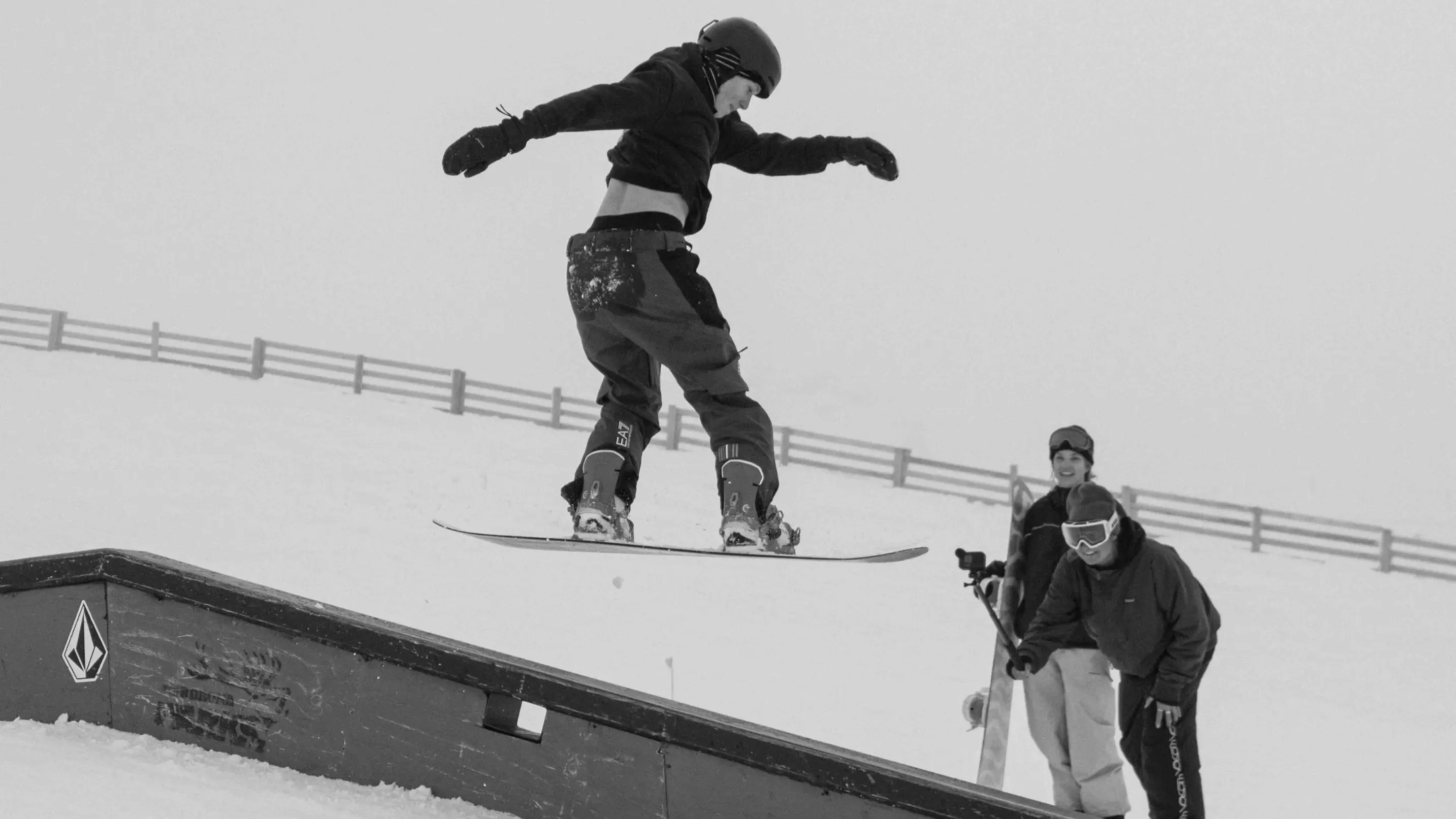 Snowboarder performs a jump onto a rail in a terrain park while two people watch and smile in the background on a snowy mountain.