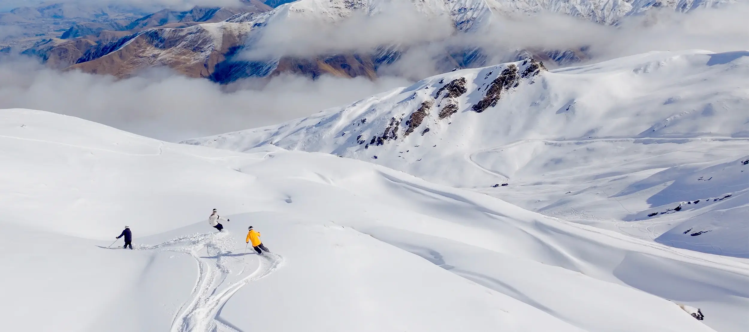 Two skiers and a snowboarder descend a wide, untouched powder slope at Cardrona, surrounded by snow-covered peaks and dramatic alpine scenery under a cloud-filled sky.