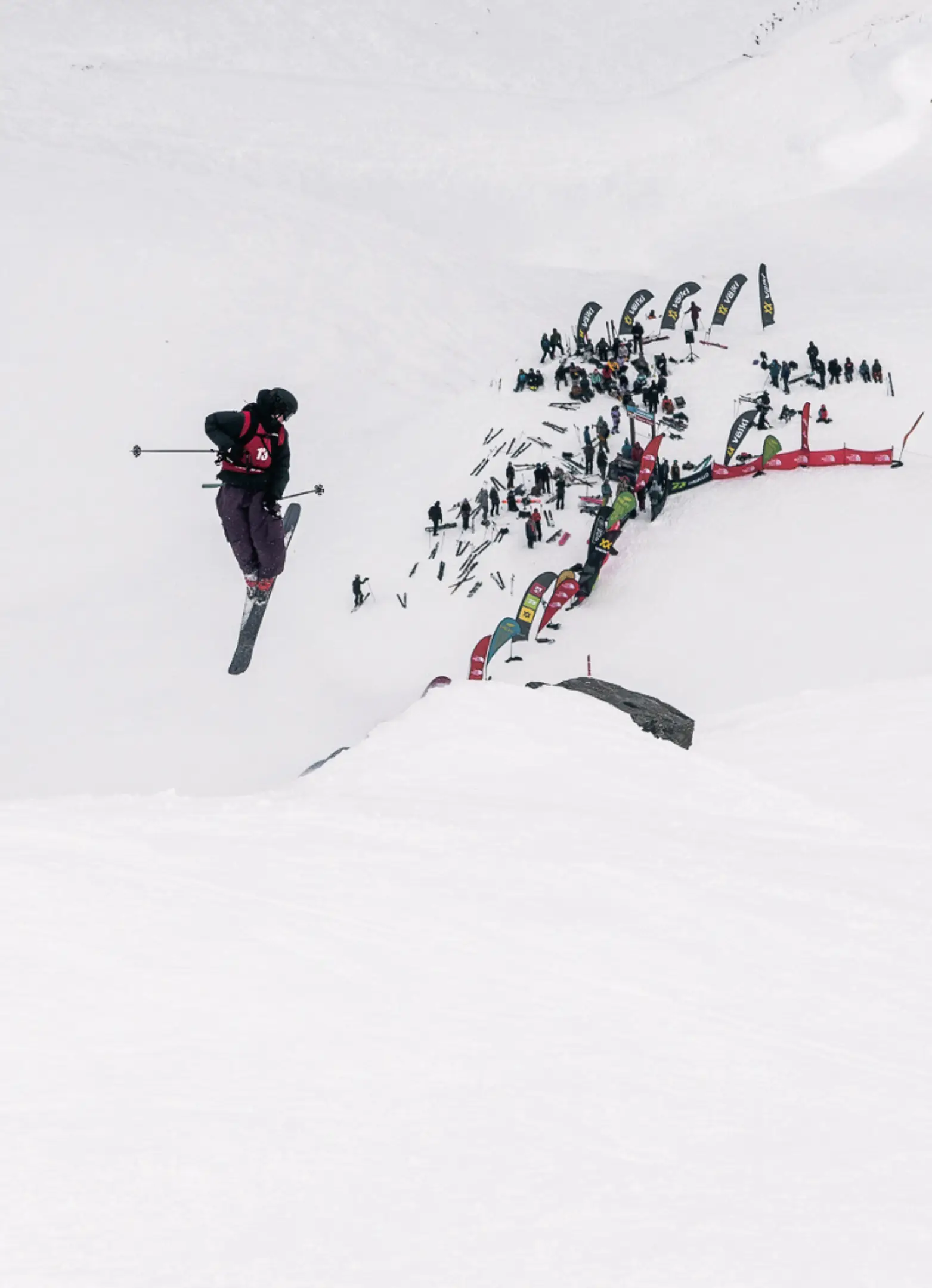 A freeskier launching off a kicker high above a crowd of spectators and sponsor flags on a snowy slope.