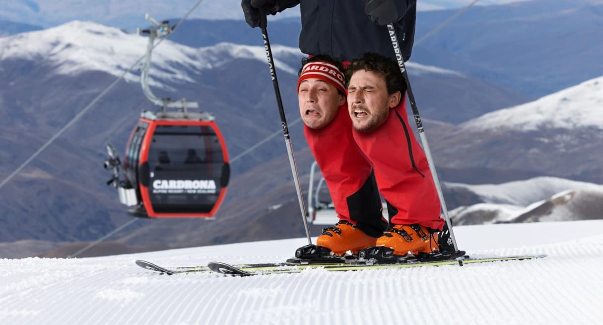 Two faces over the knees of a skier on a groomed slope with mountain scenery and a Cardrona branded gondola in the background.