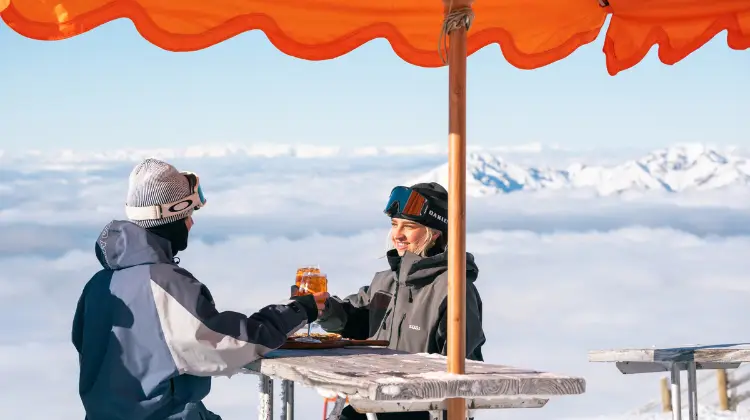 Two friends cheers an aperol sitting at an outdoor table in an alpine environment, dressed in ski clothing.