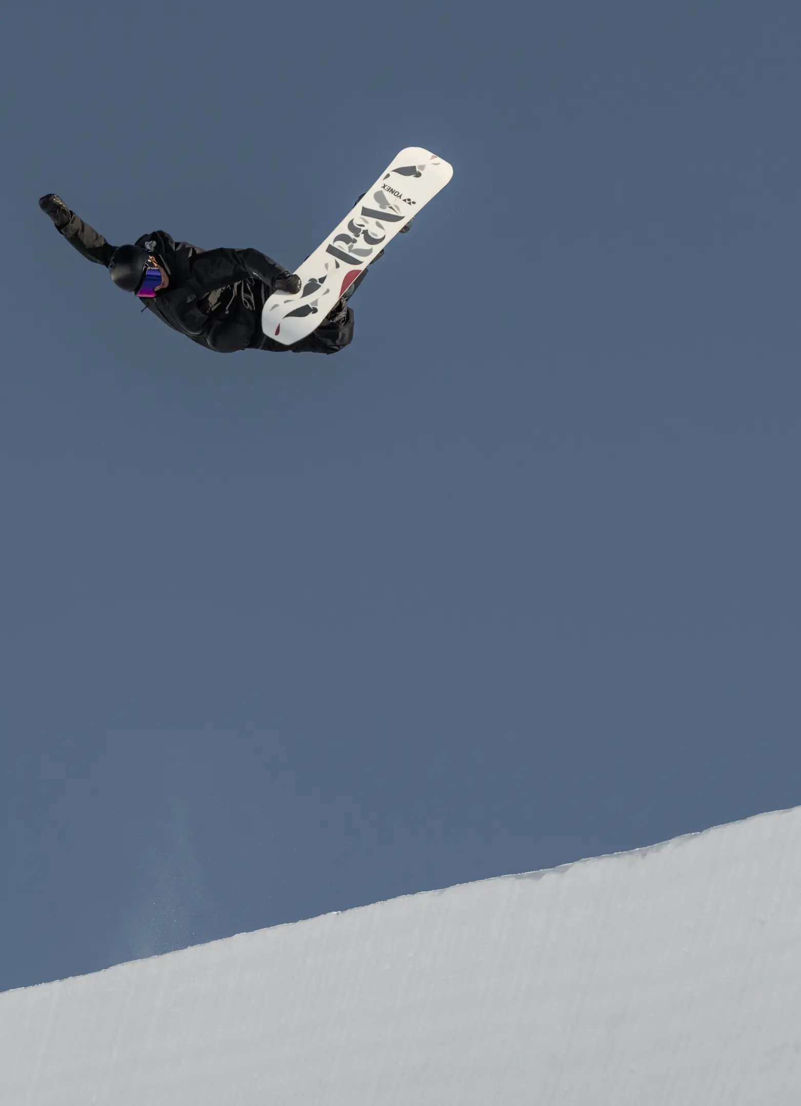 A snowboarder performs an inverted trick high above the halfpipe lip against a clear blue sky.