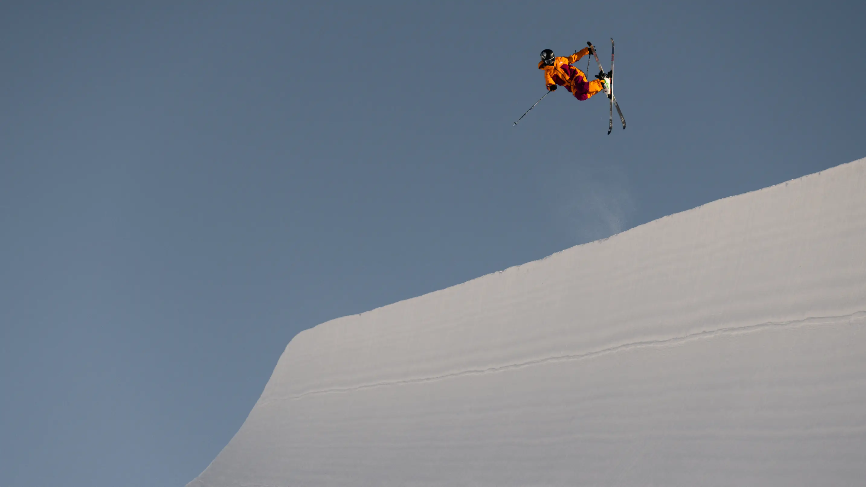 A freestyle skier in a bright orange jacket crosses their skis mid-air above the halfpipe lip against a clear blue sky.