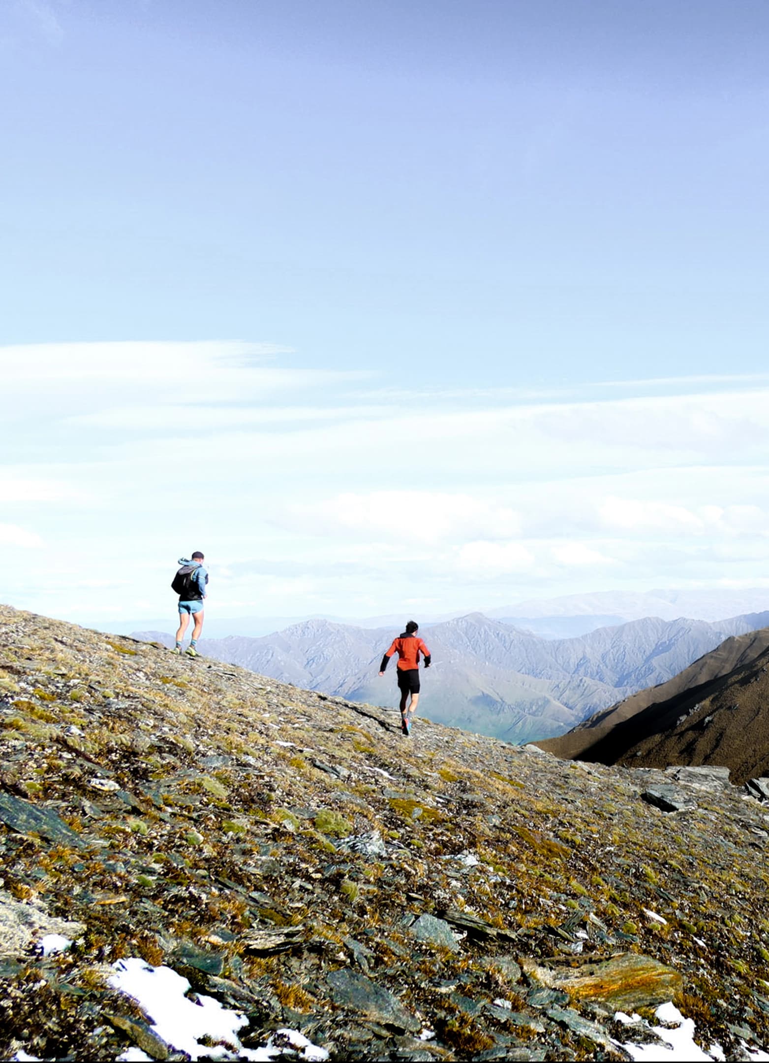 Two people running along the ridge of a rocky mountain on a clear blue skies day.