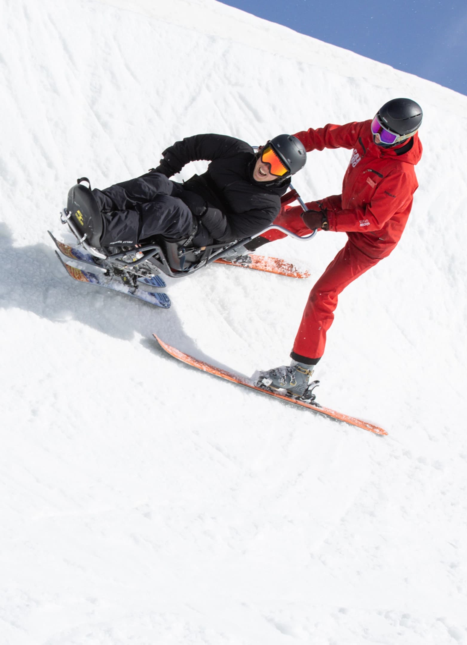 A sit-ski rider is guided downhill by an instructor in red on a snowy slope.