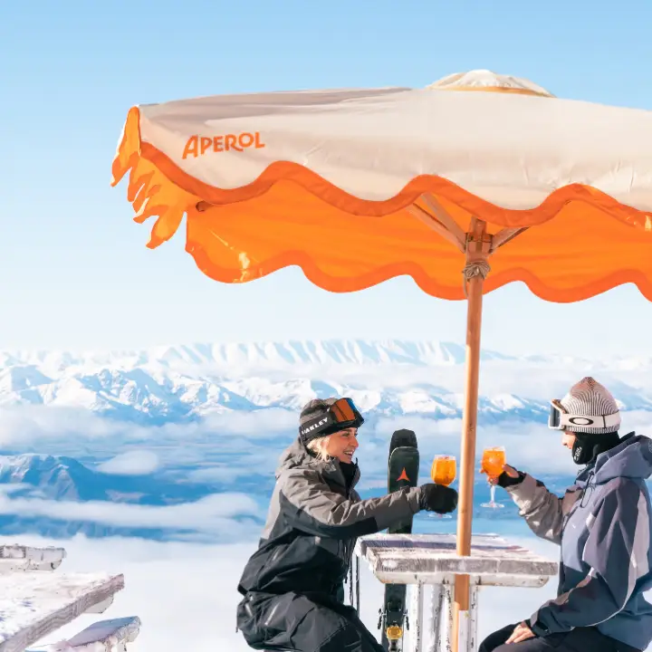 Two skiers clink Aperol Spritz glasses at a mountaintop bar table, snow-capped peaks and clouds behind them.