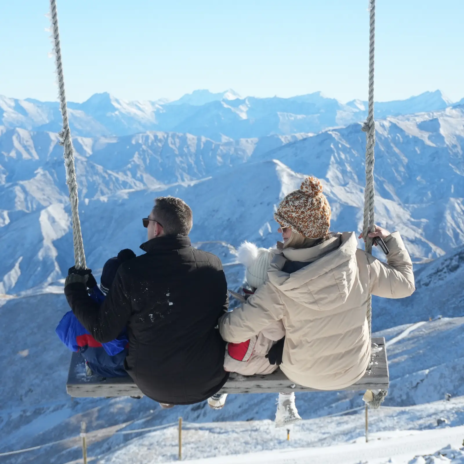 A family of four sit on the famous Cardrona swing, found at the top of the McDougall's chairlift, and look out at the snowy Southern Alps on a clear blue sky day.