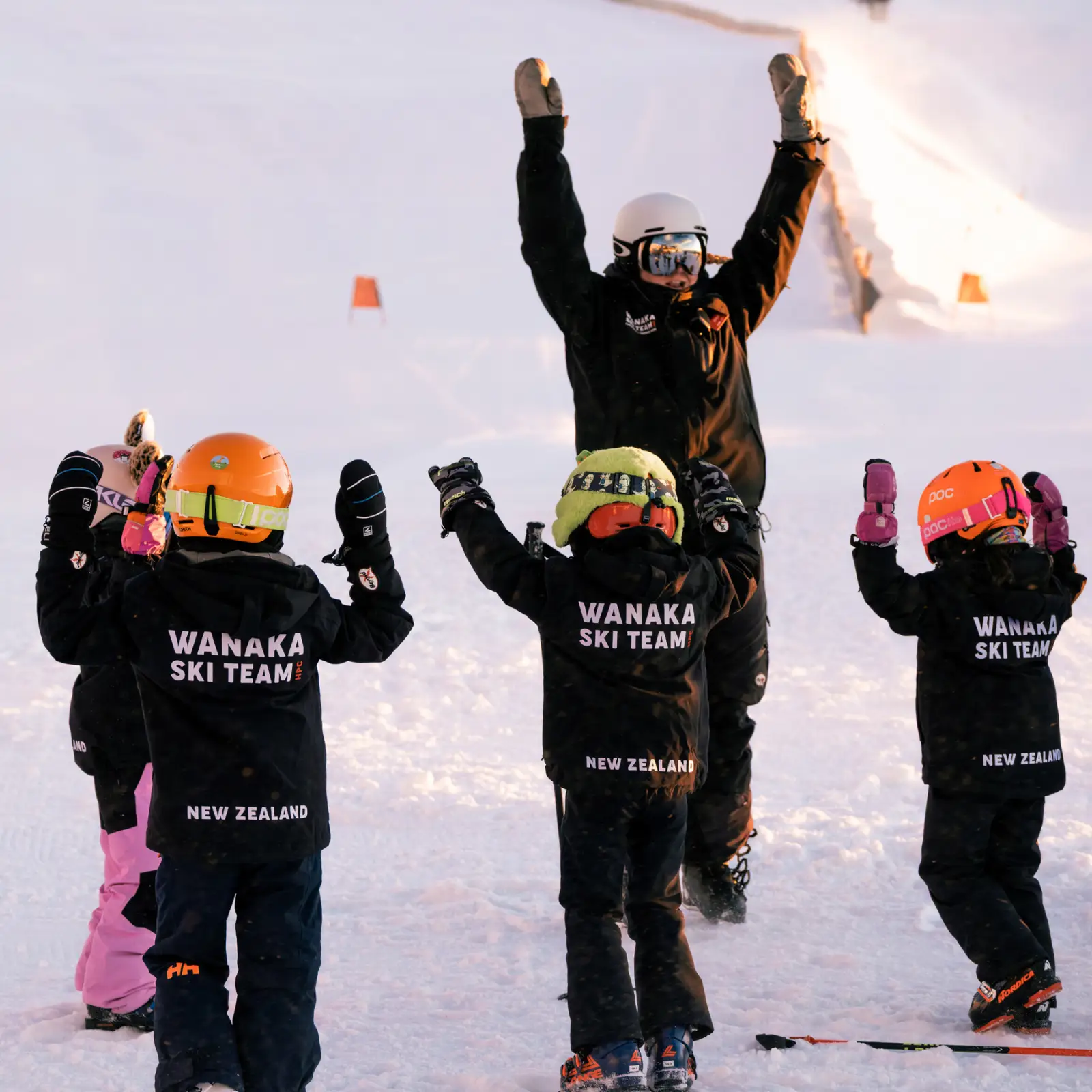 An instructur leads a group of young skiers in a warm up exercise, all wearing black Wanaka Ski Team branded jackets.