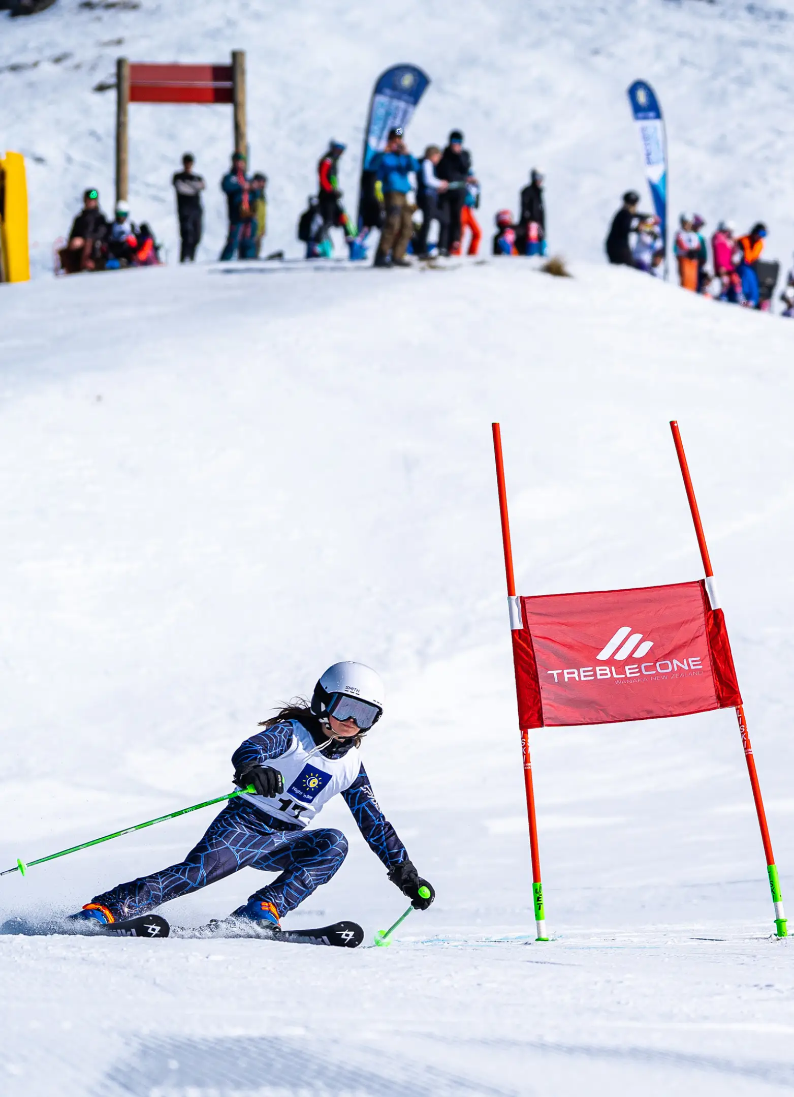 A young female ski racer carves through red Treble Cone branded gates during a giant slalom race, with a crowd of spectators watching from the snowy hillside above.