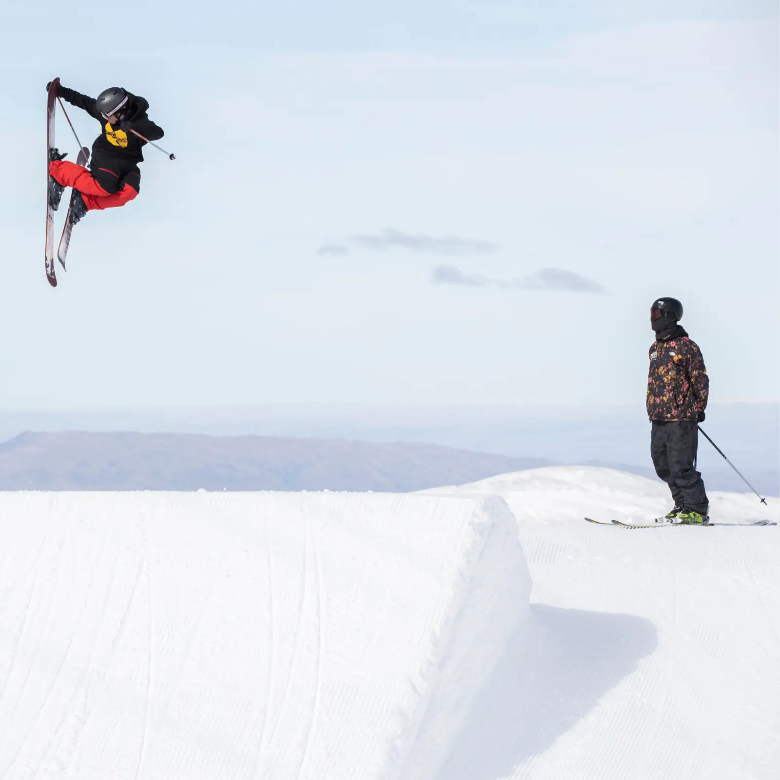 A skier in red and black throws a big inverted trick off a park jump while a coach watches from the landing zone, with hazy mountains visible in the background.